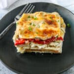 A slice of vegetable lasagna with white sauce on a black plate.