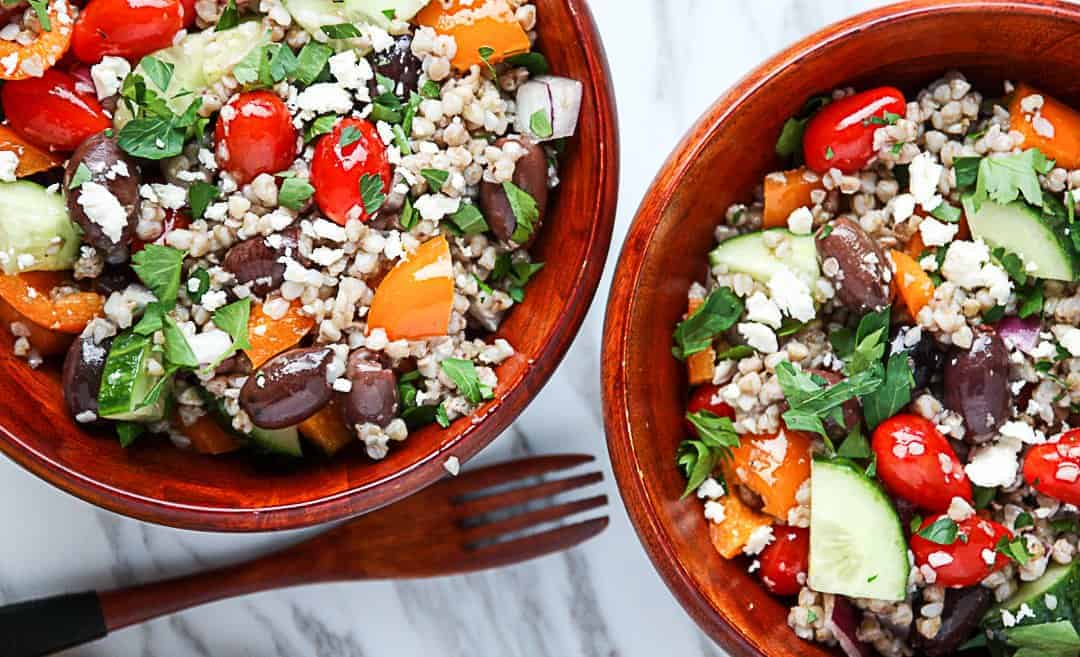 Two wooden bowls of salad with a wooden fork on a marble table.
