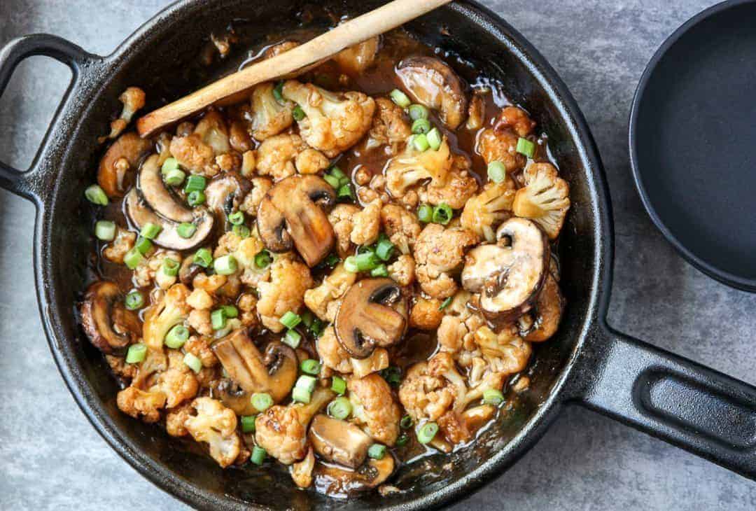 A skillet with mushrooms and cauliflower is stirred with  a wooden spoon