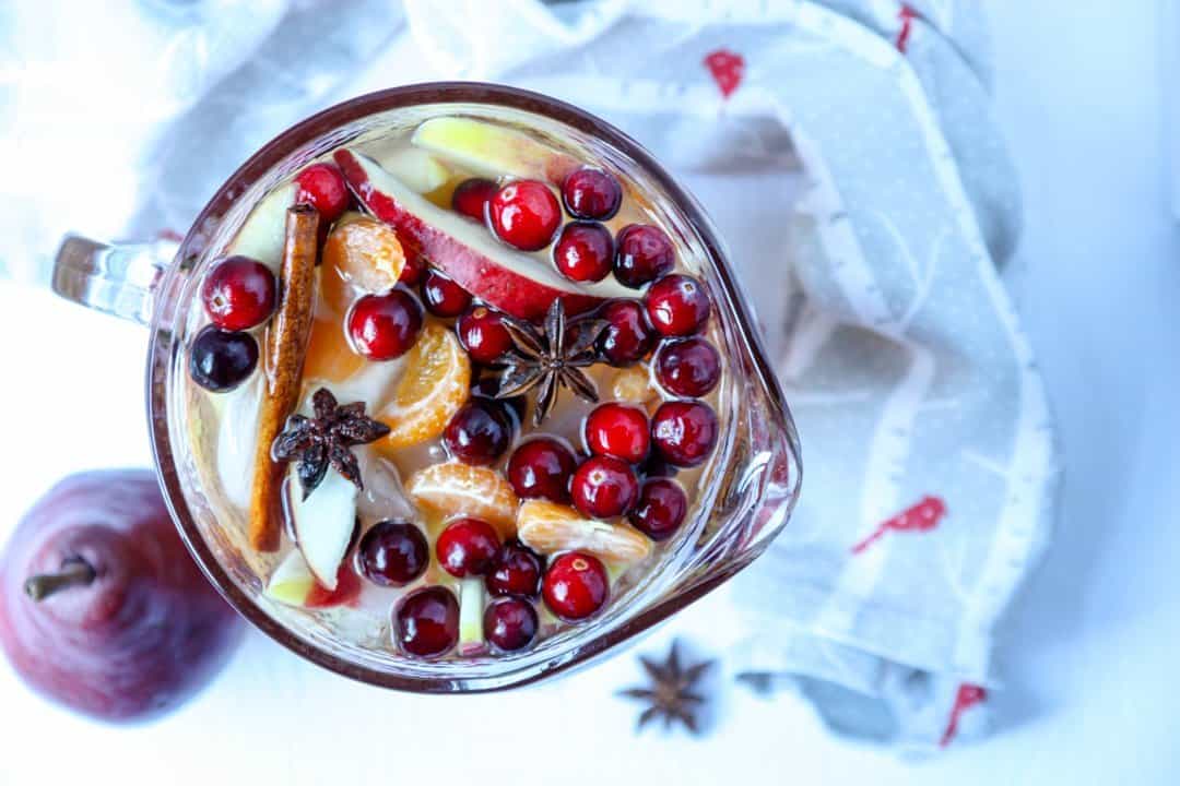 Top down view of carafe of sangria filled with cinnamon sticks, apple slices, orange slices, and cranberries