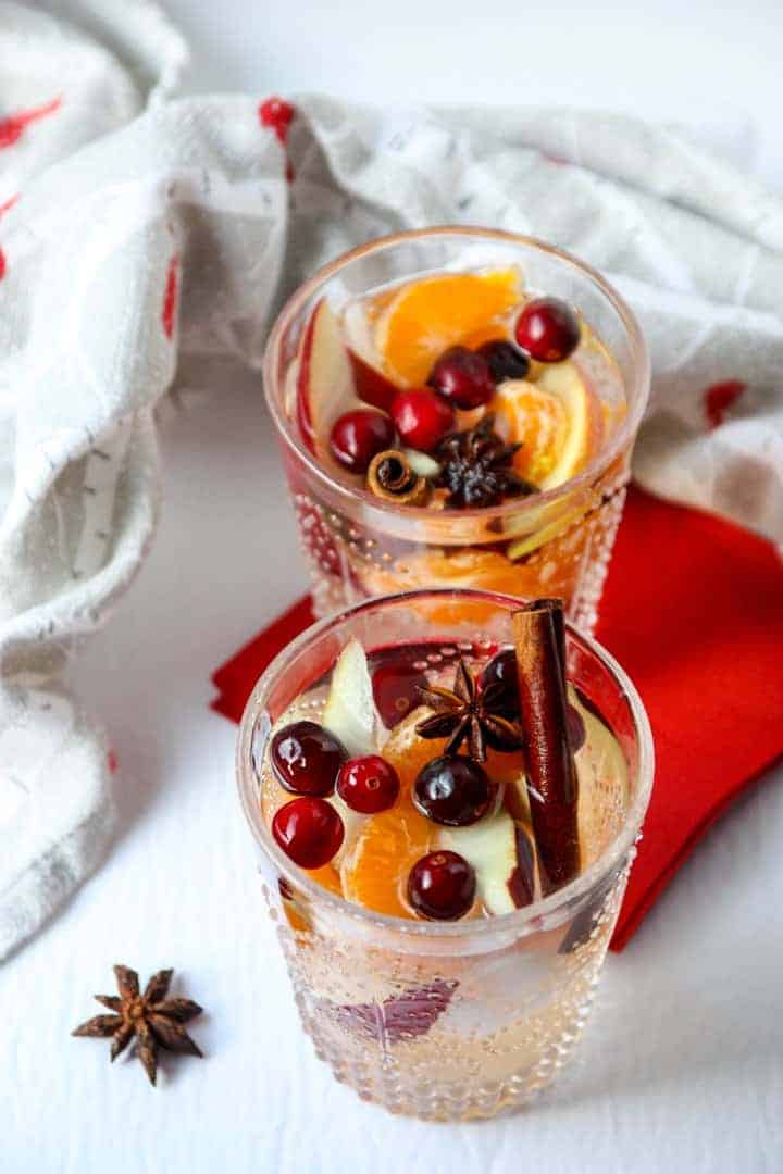 Two glasses of white sangria filled with cinnamon sticks, apple slices, orange slices, and cranberries