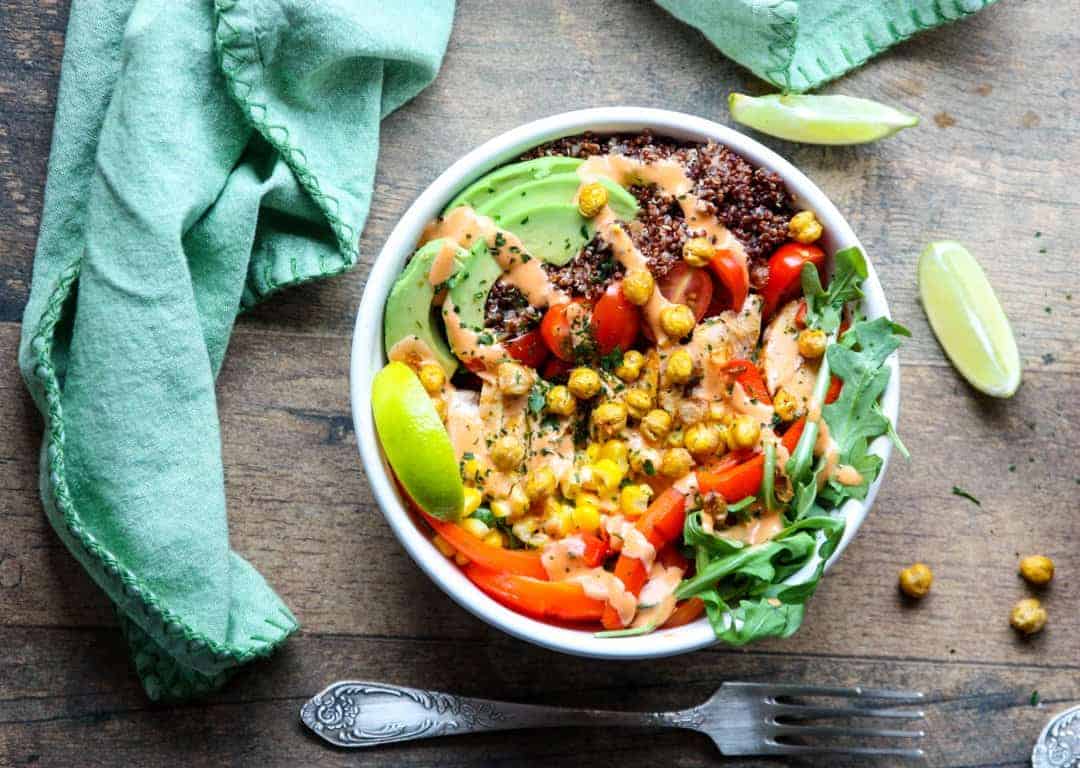 A bowl of quinoa topped with vegetables, avocado and sauce on a wooden table.