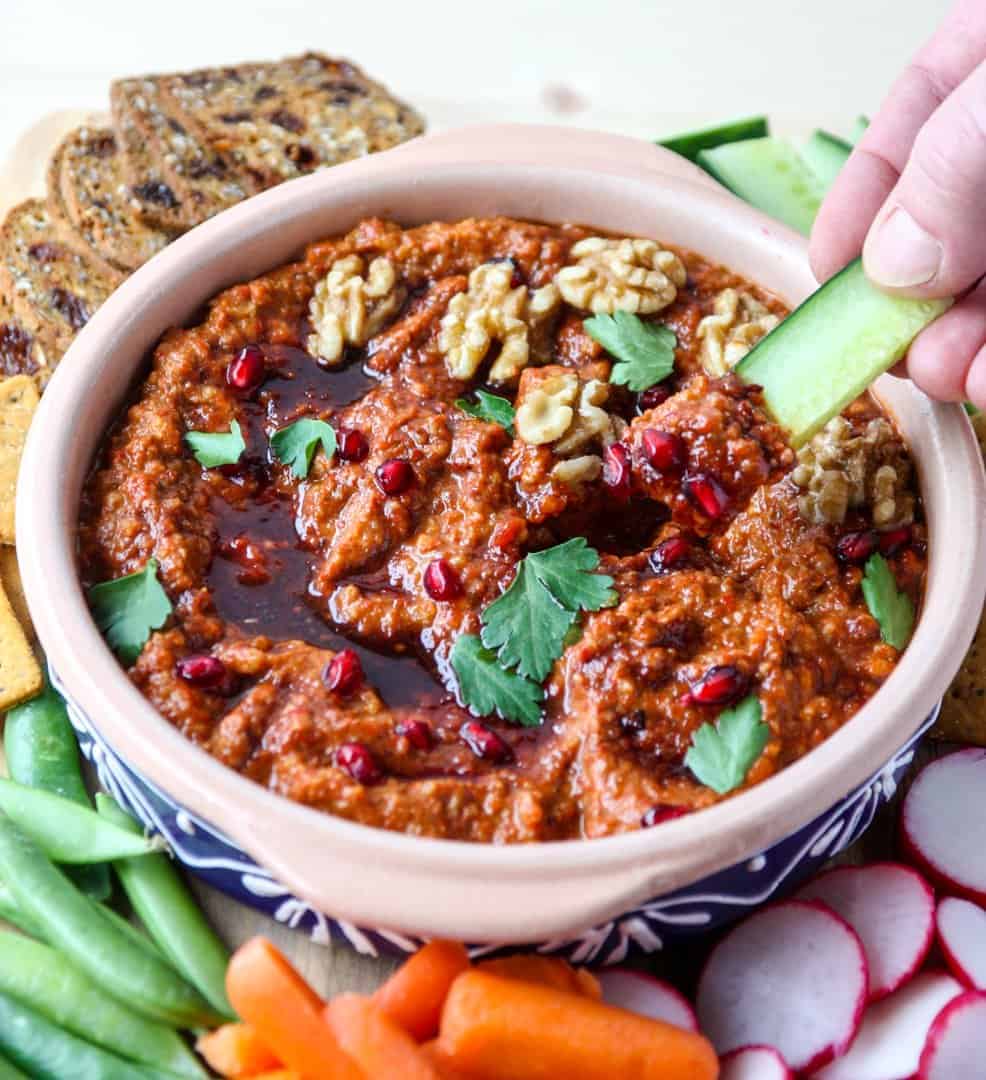 A bowl of dip with vegetables and cracker with fingers dipping a celery stick
