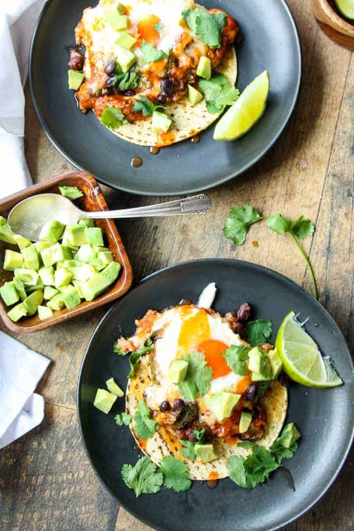 Two plates of huevos ranceheros served on corn tortillas on black plates.