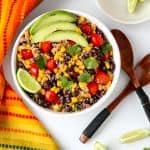 A bowl of salad on a white table with two wooden salad spoons and a small dish of lime wedges.