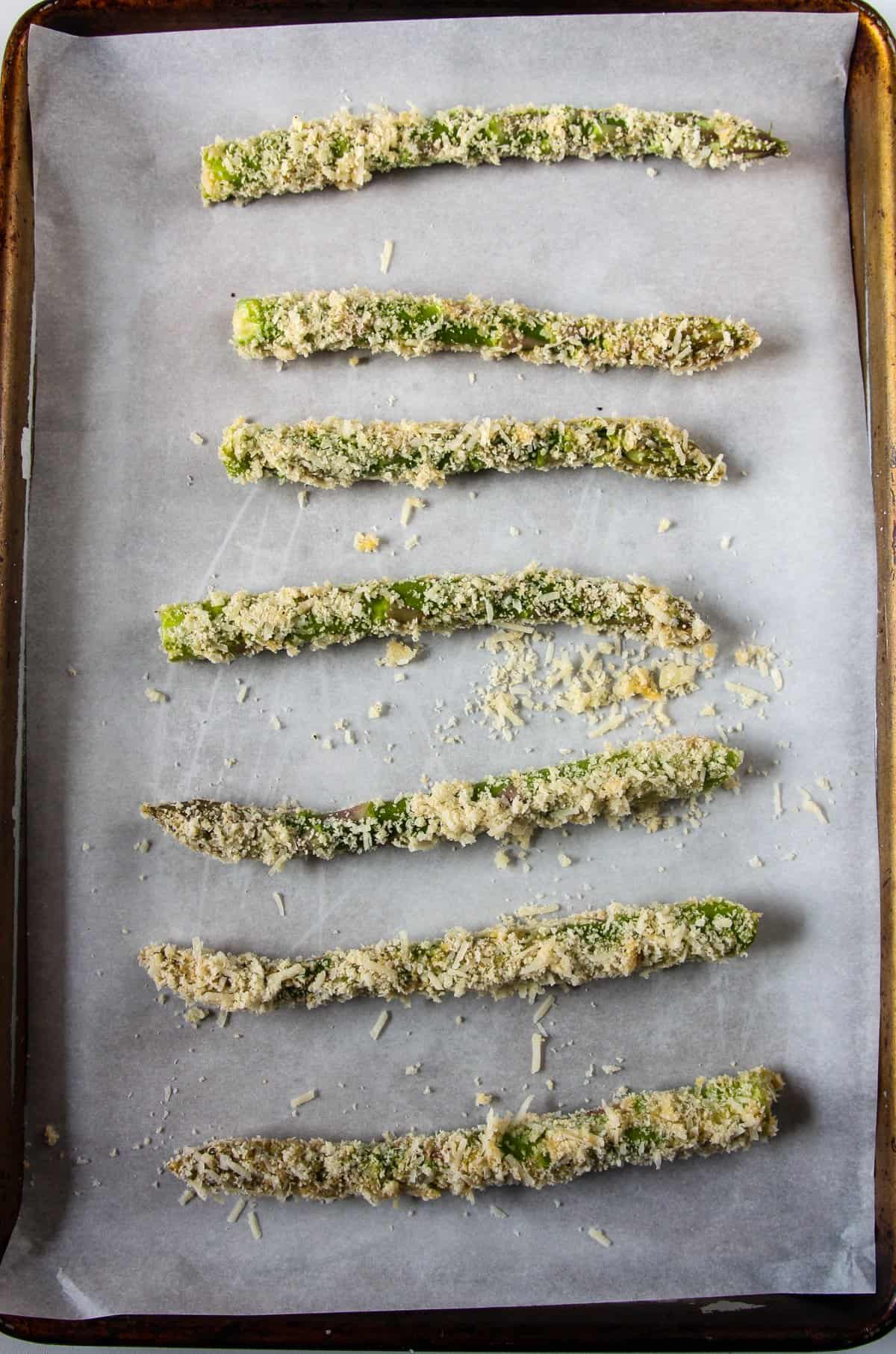 Coated asparagus fries on a parchment lined baking sheet ready to go into the oven.