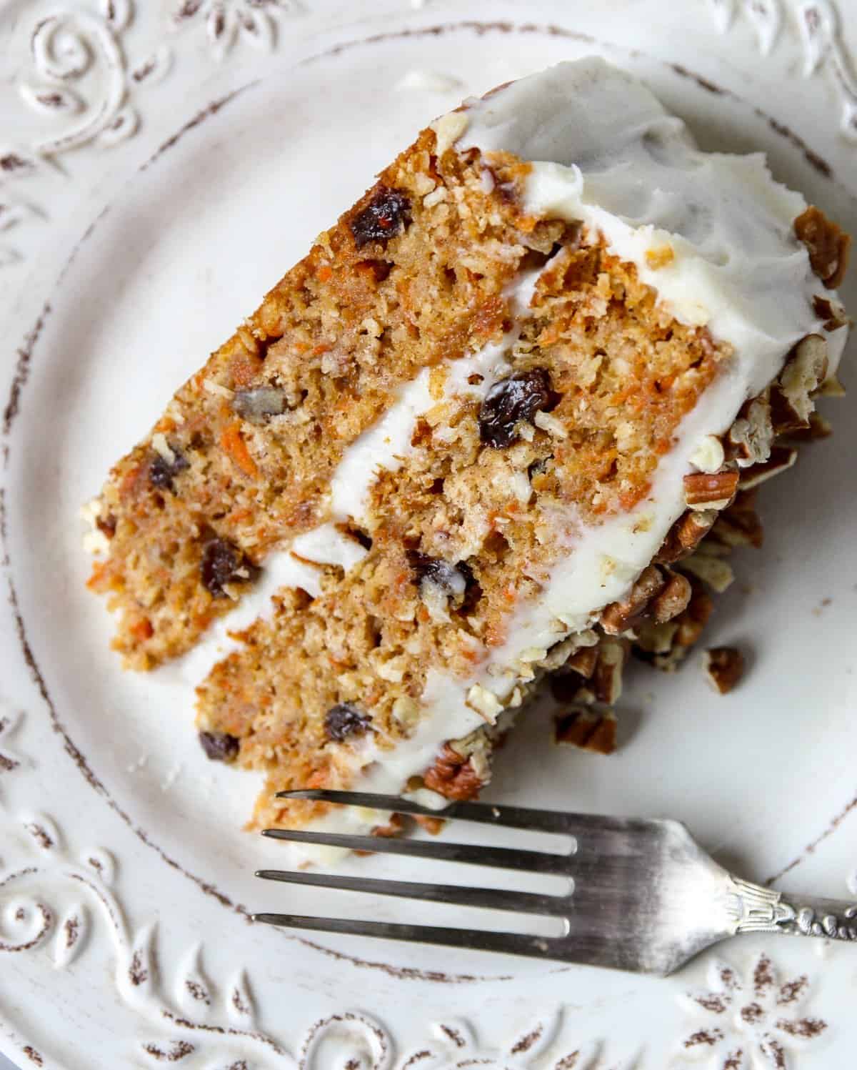 A slice of frosted carrot cake on a white plate with a silver fork.