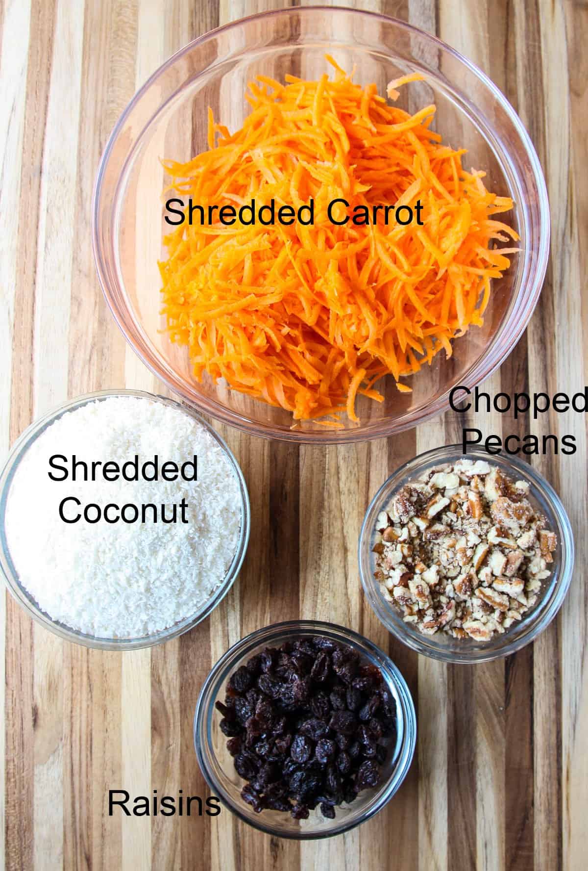 Four glass bowls containing grated carrot, coconut, nuts, and raisins.