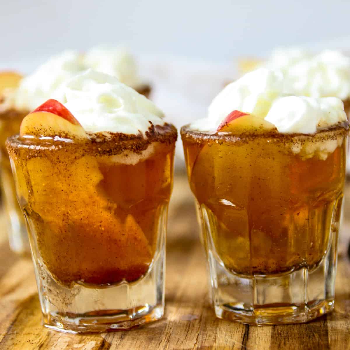 Two shot glasses with apple pie shooters and topped with whipped cream.