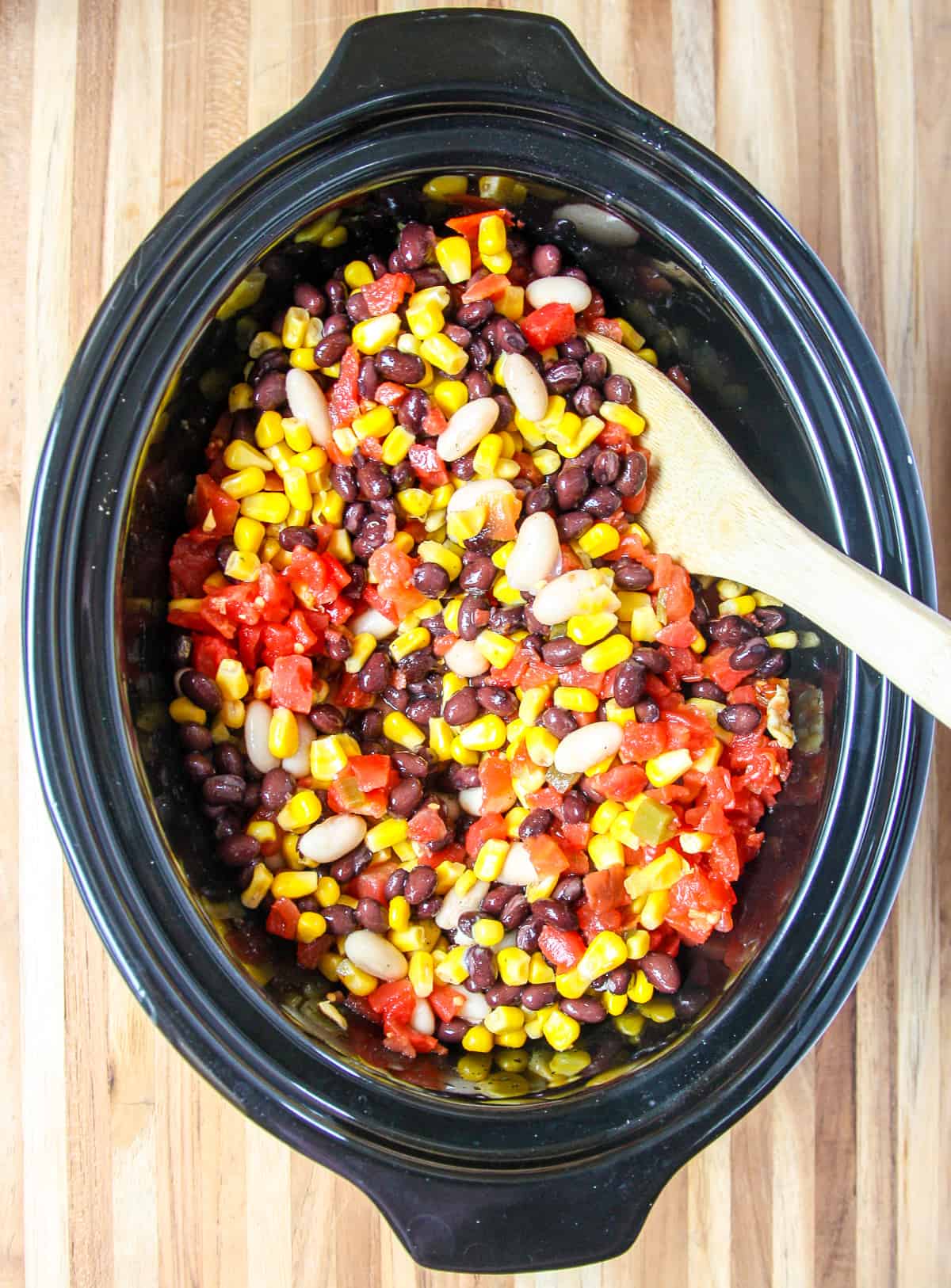 The beans, corn, and tomatoes being stirred into the slow cooker with a wooden spoon.