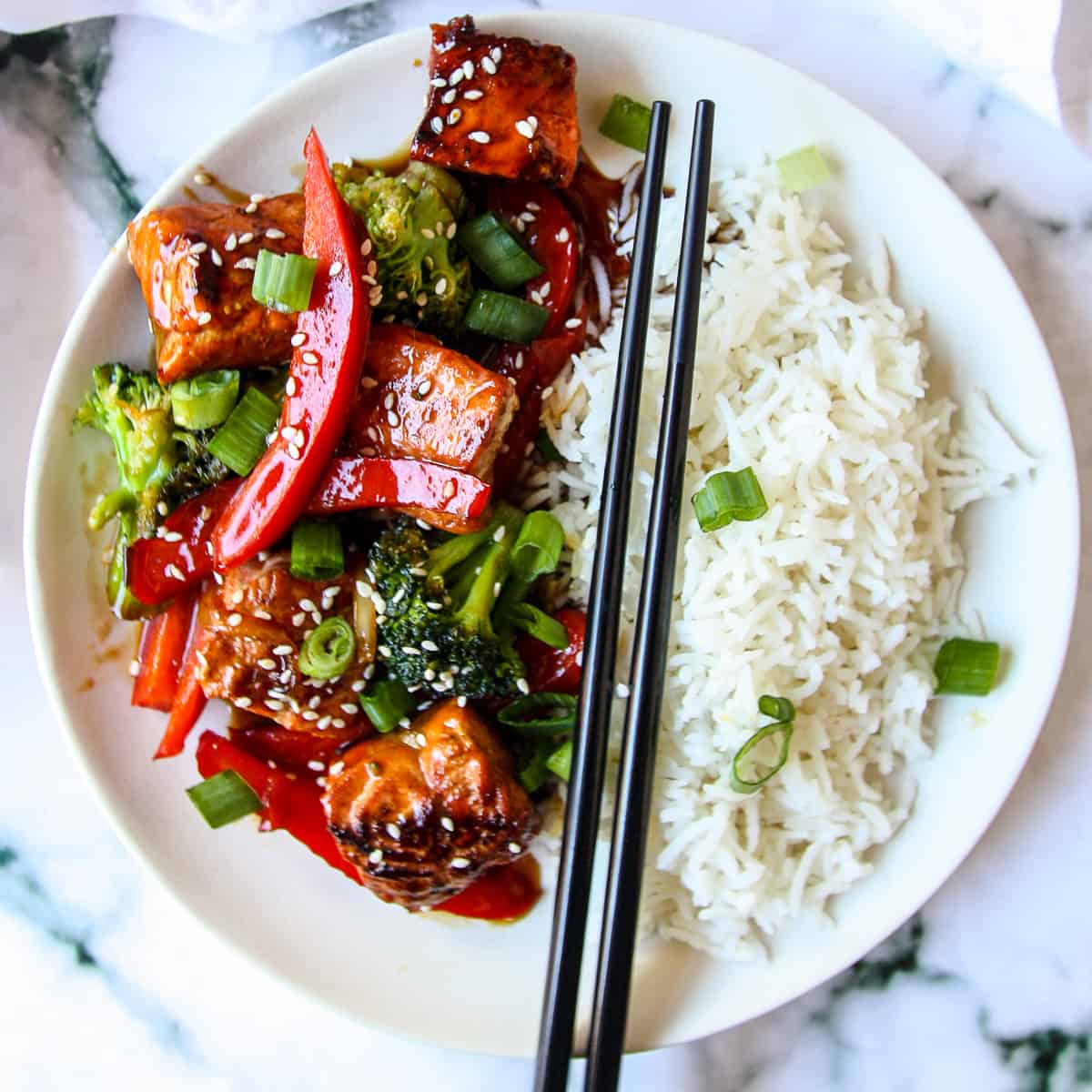 Salmon stir fry and a side of rice on a white plate with black chopsticks.