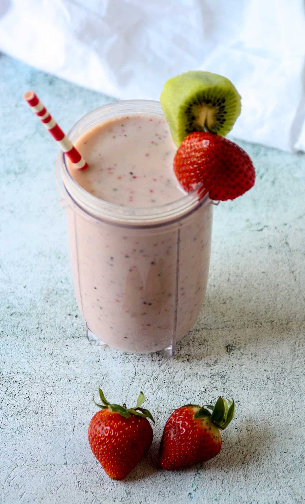 A tall glass of kiwi and strawberry smoothie on a white textured surface next to two whole strawberries.