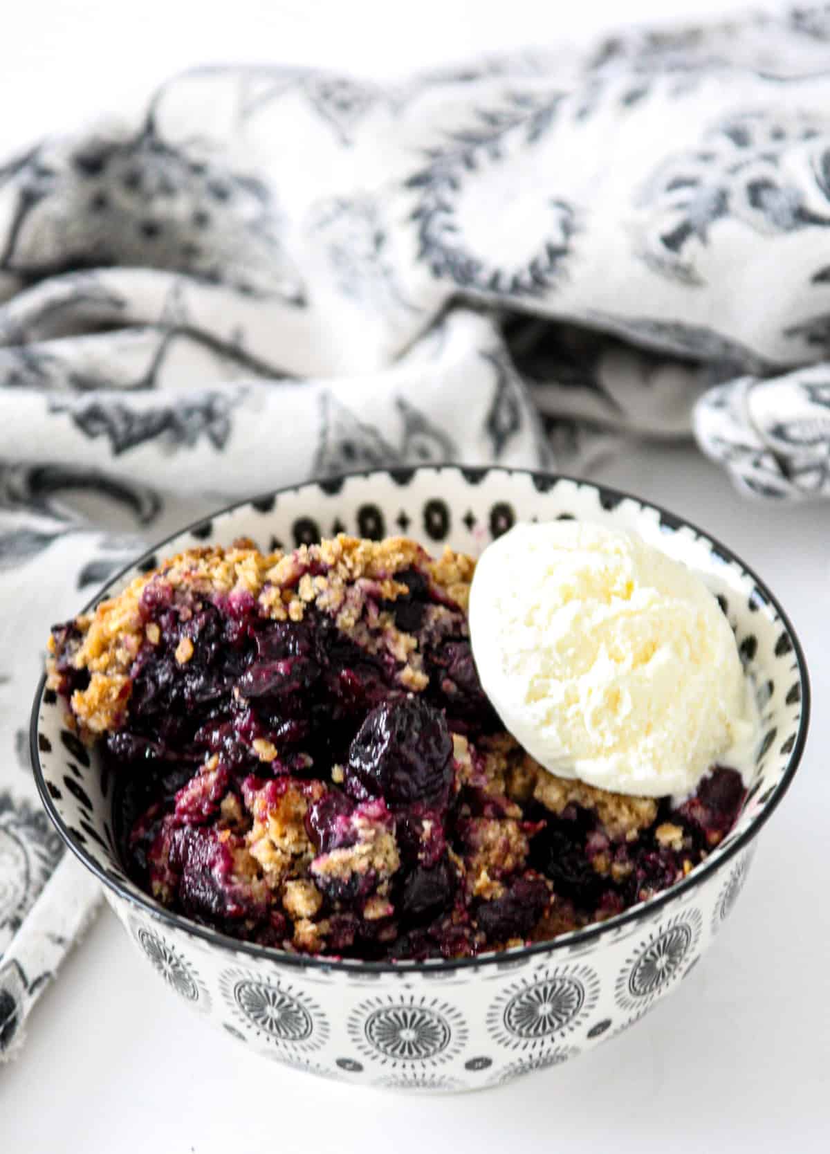 Gluten free blueberry crisp in a black and white bowl, topped with a scoop of ice cream.