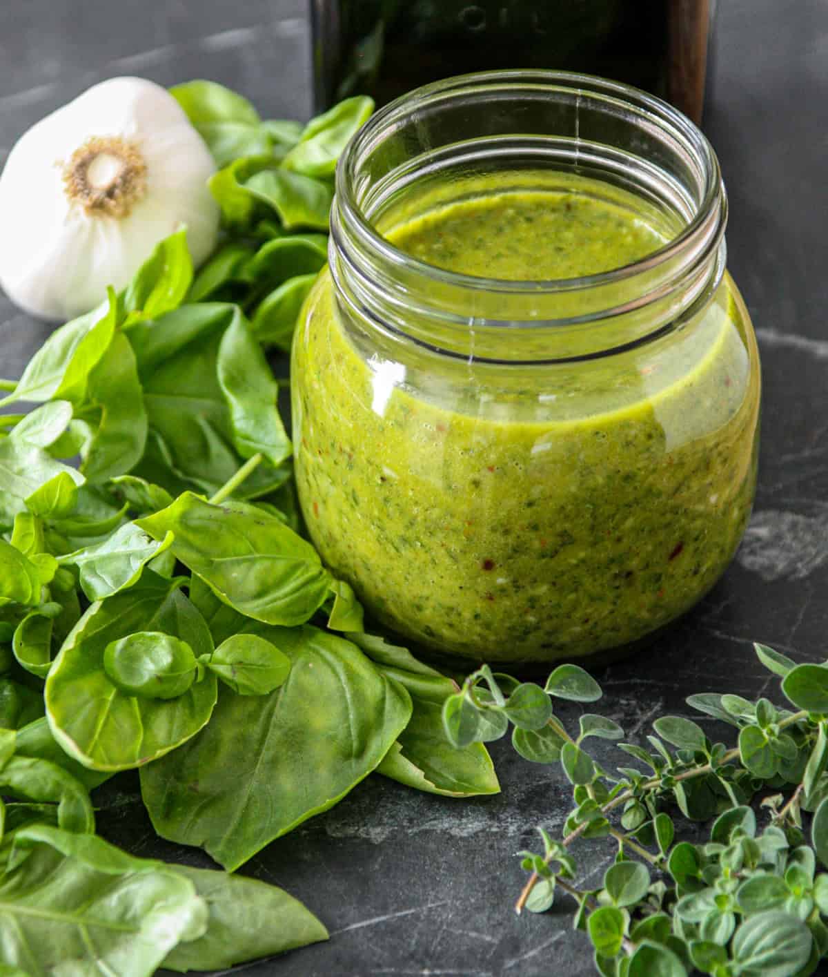 Basil chimichurri sauce in a glass jar, surrounded by fresh herbs.